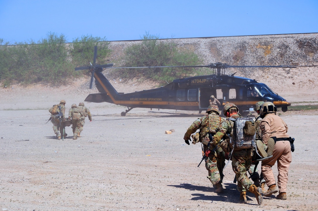Soldiers and U.S. Customs and Border Protection personnel carrying two stretchers with objects on them run toward a helicopter on sandy terrain, with a hill and blue sky in the background.
