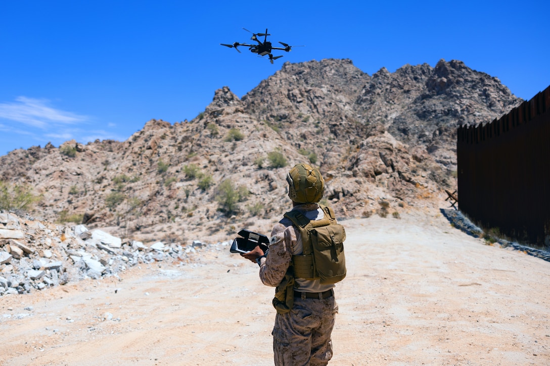 A Marine standing in a sparsely vegetated area near a tall security wall operates a small drone hovering above, with a hill and blue sky in the background.