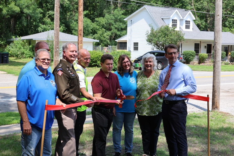 Group of 8 people behind a red ribbon. Scissors are used to cut the ribbon while group smiles at the cameras.