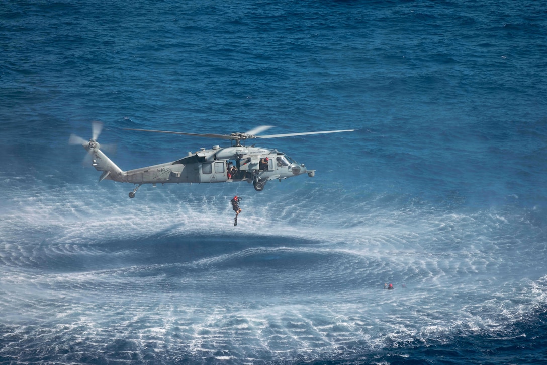 A sailor wearing swimming gear jumps out of a helicopter as it hovers over a body of water.