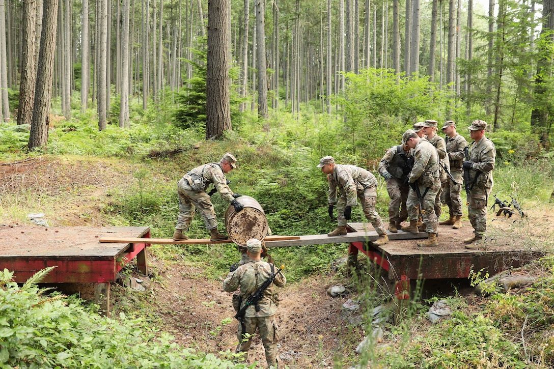 Army officer candidates roll a barrel over a wooden plank in a wooded area.