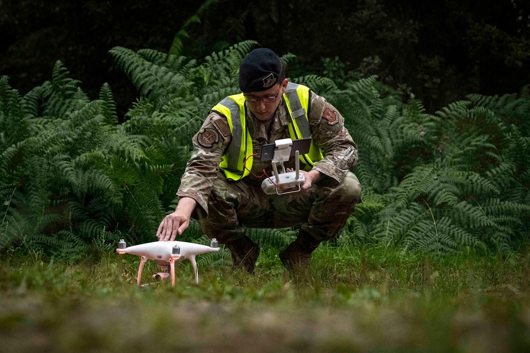 A squatting airman holds a remote in one hand while tapping a drone in the other in a wooded area.