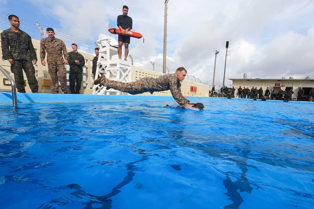 A Marine holding a pack dives into a pool as fellow service members watch from the sideline.