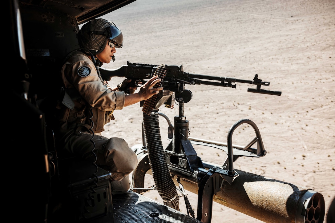 A kneeling Marine fires a weapon from an airborne aircraft over a desert-like area.