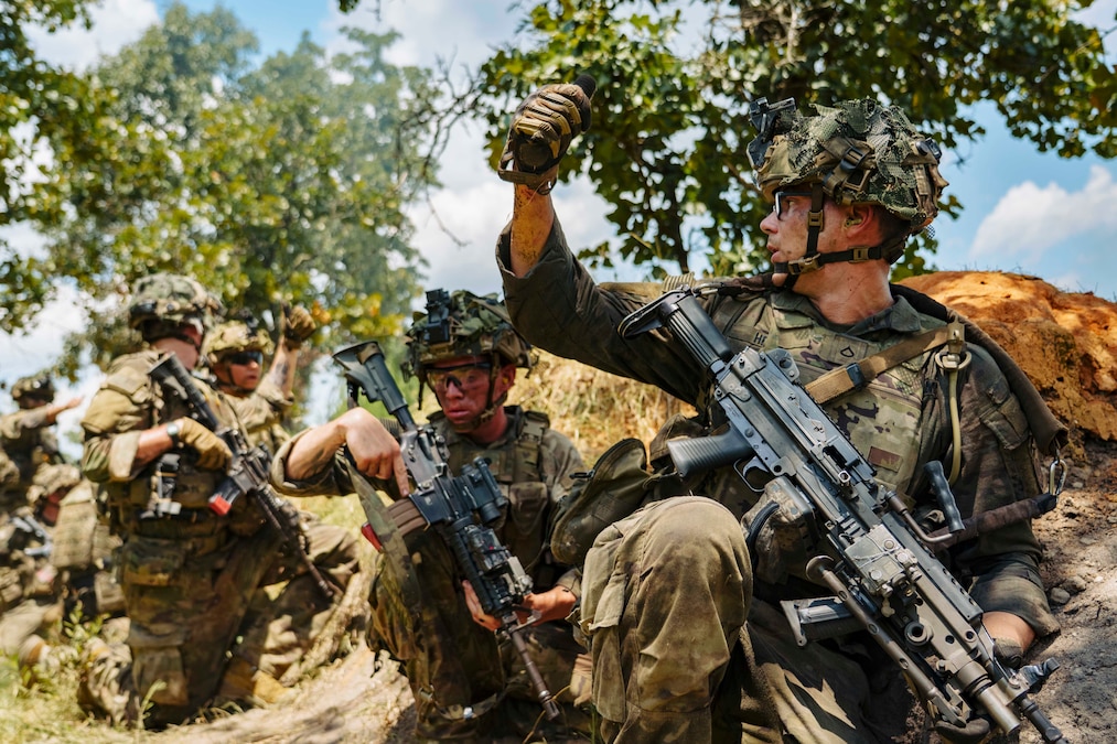 Soldiers in tactical gear communicate with hand signals while kneeling in a wooded area during the day.