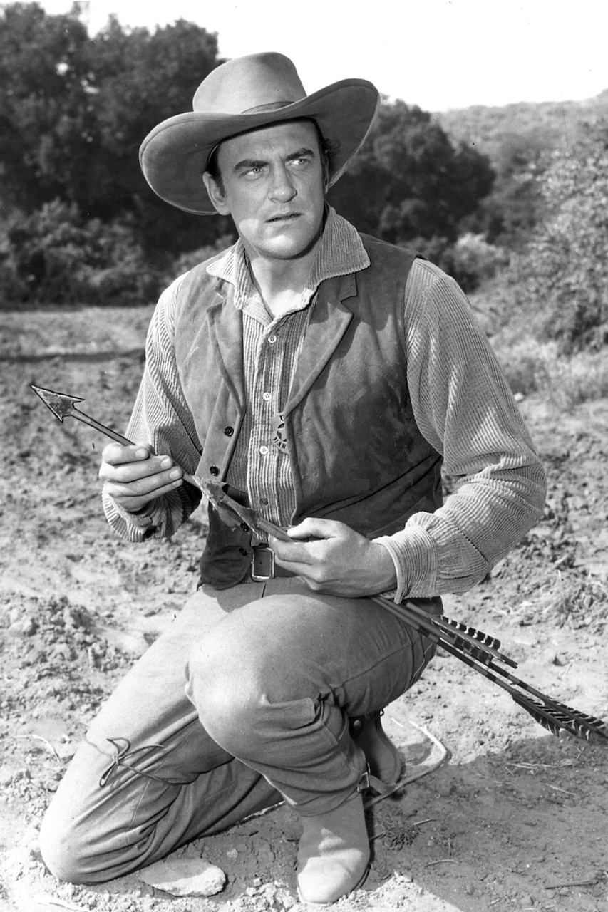 A man in a cowboy costume holds arrows and stares into the distance.