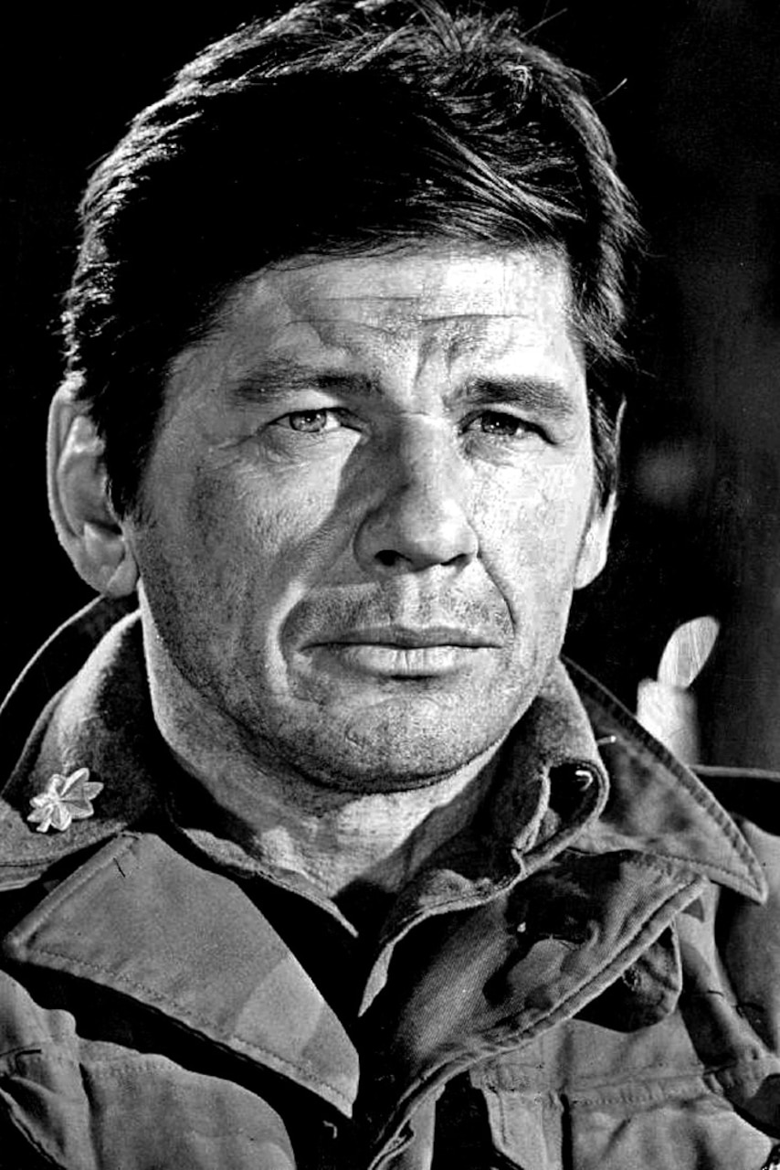 A man in uniform without a head cover on looks into the distance, against a dark background.