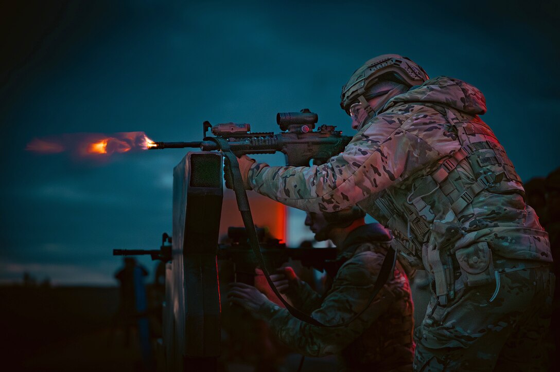 An airman fires a weapon at an outdoor range, creating a flash against a dark blue sky.