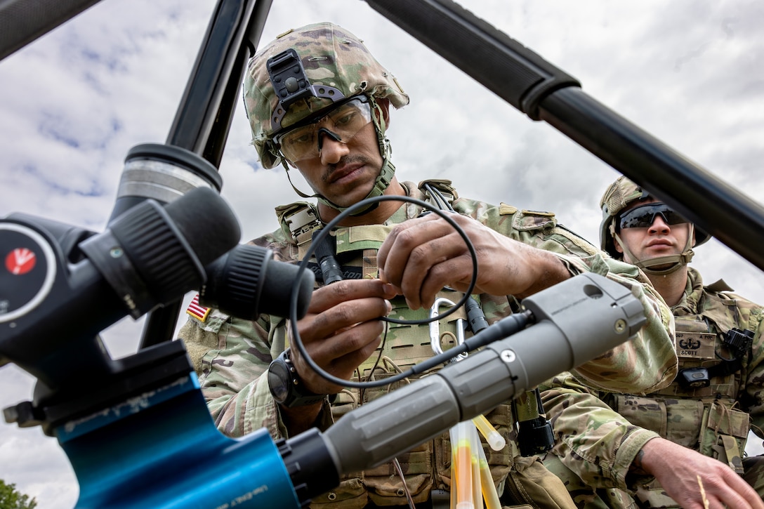 A soldier assembles a device comprising rods and cables outdoors as another soldier looks on.