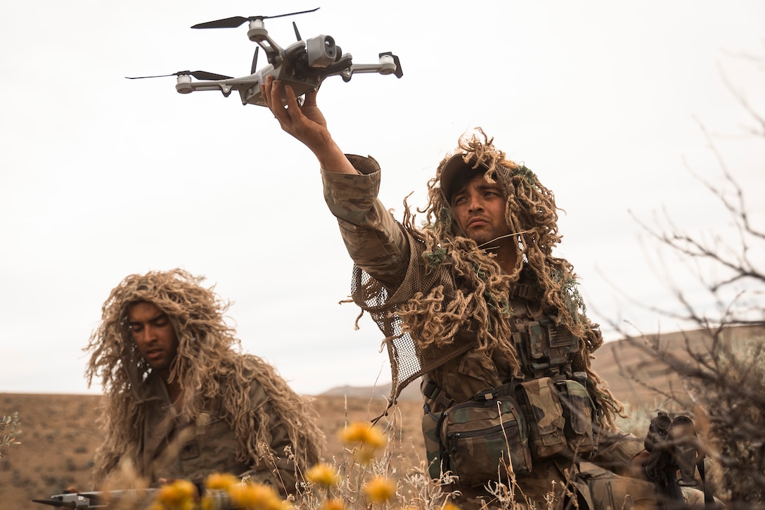 A soldier wearing ghillie suit-type camouflage holds up a drone in a field as another soldier sits nearby.