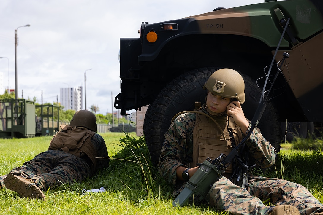 Two Marines hide behind a vehicle, one talks on a radio.