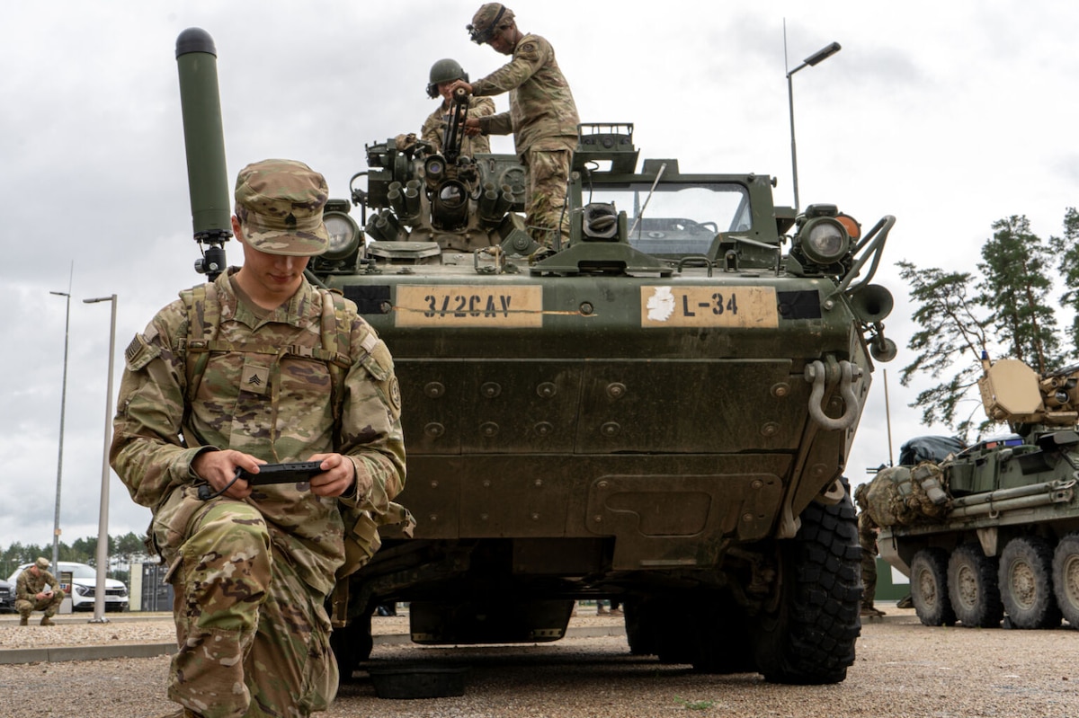 A uniformed service member takes a knee and operates a handheld device. Behind him is a combat vehicle with two other service members standing on top.