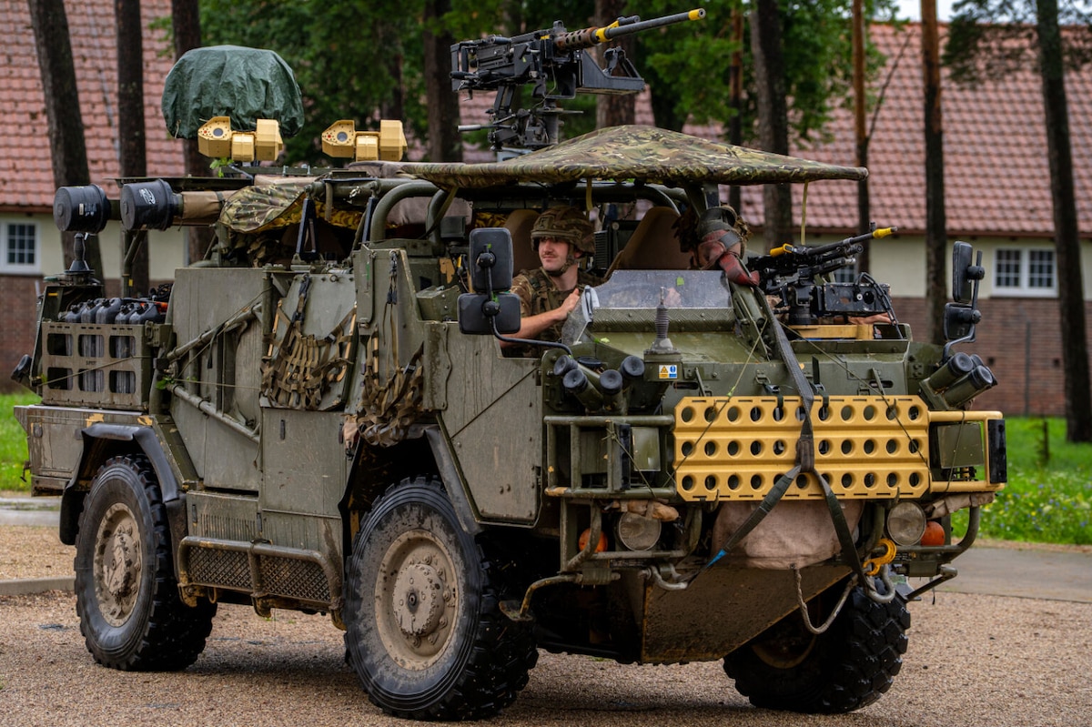 A combat vehicle rolls across a gravel road. The vehicle has a gun mounted on top and camouflage material over the heads of the driver and passenger.