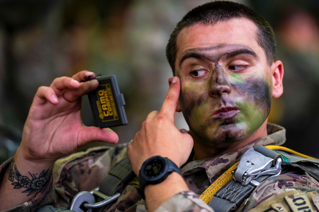 A soldier applies face paint while looking at a compact mirror against a blurred background.