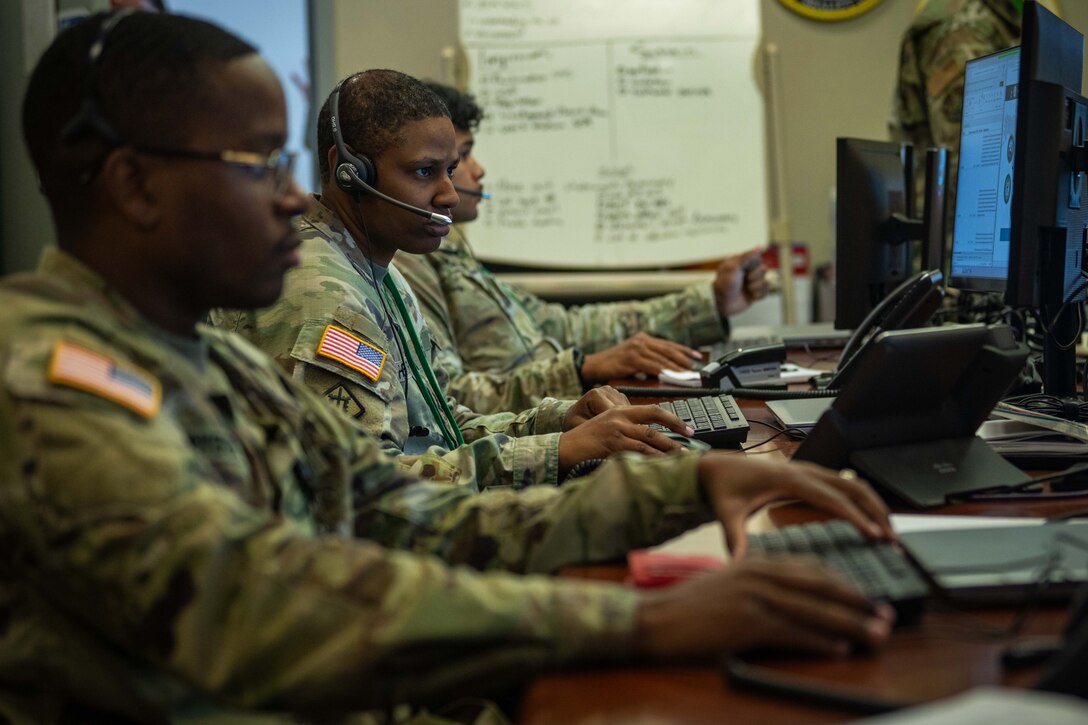 Service members wearing headsets and seated at a large desk with  computer keyboards and monitors work in an office environment.