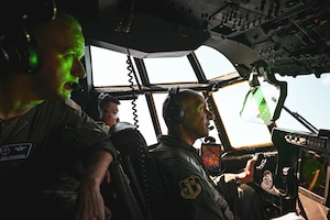 Men in uniform stand in the flight deck of an aircraft while it is being flown.