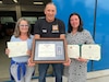 Thomas Moosmann, the supervisor at the Privately Owned Vehicle (POV) inspection station and the Base Support Operations Maintenance facility in Baumholder, Germany, (center) and his wife, Silke Moosmann, (left) received multiple certificates of achievement and tokens of appreciation July 31, 2025, for his nearly 45 years of service with the U.S. Army as a German host nation employee. (Photo by Stefan Pfister)