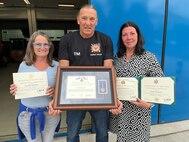 Thomas Moosmann, the supervisor at the Privately Owned Vehicle (POV) inspection station and the Base Support Operations Maintenance facility in Baumholder, Germany, (center) and his wife, Silke Moosmann, (left) received multiple certificates of achievement and tokens of appreciation July 31, 2025, for his nearly 45 years of service with the U.S. Army as a German host nation employee. (Photo by Stefan Pfister)