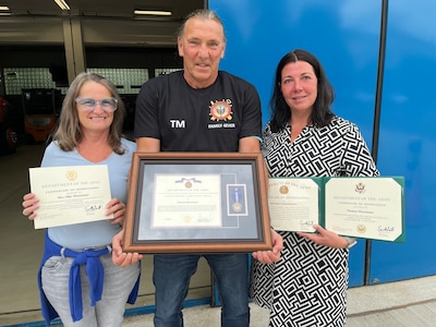 Thomas Moosmann, the supervisor at the Privately Owned Vehicle (POV) inspection station and the Base Support Operations Maintenance facility in Baumholder, Germany, (center) and his wife, Silke Moosmann, (left) received multiple certificates of achievement and tokens of appreciation July 31, 2025, for his nearly 45 years of service with the U.S. Army as a German host nation employee. (Photo by Stefan Pfister)