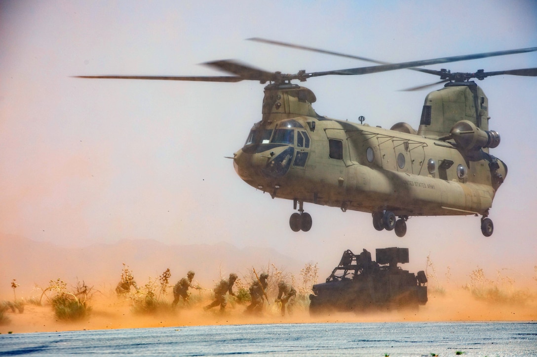 Soldiers work to attach a cable to a Humvee as a helicopter hovers above the vehicle, kicking up dust in a sparsely vegetated area.