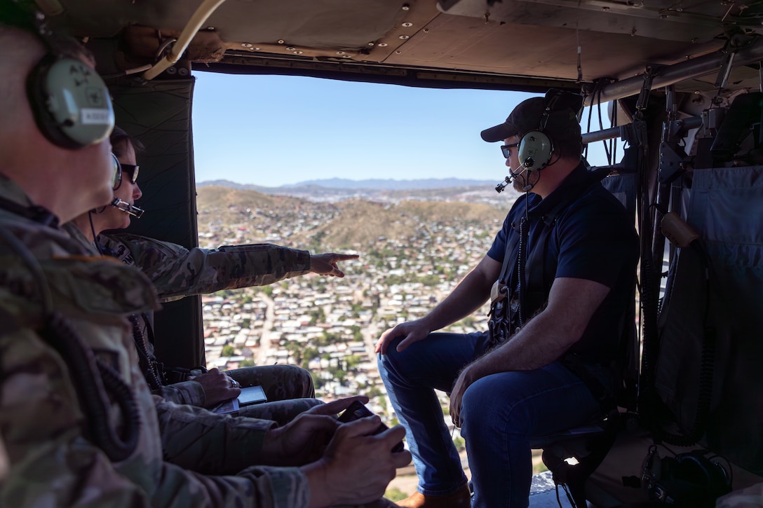 A soldier sitting next to another soldier points out an open door of an airborne helicopter as another person sitting across from the soldiers looks at the community below.