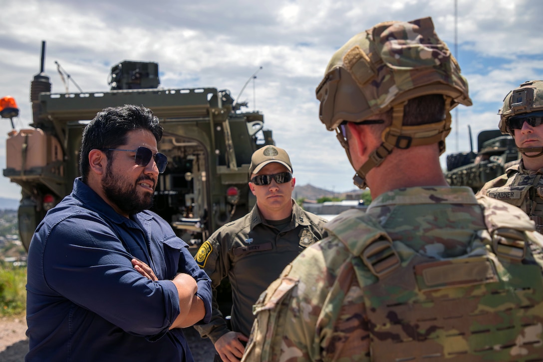 A person wearing a blue shirt and sunglasses talks to a soldier wearing tactical gear as another soldier and a person wearing a green cap and shirt stand nearby, with an armored vehicle in the background.
