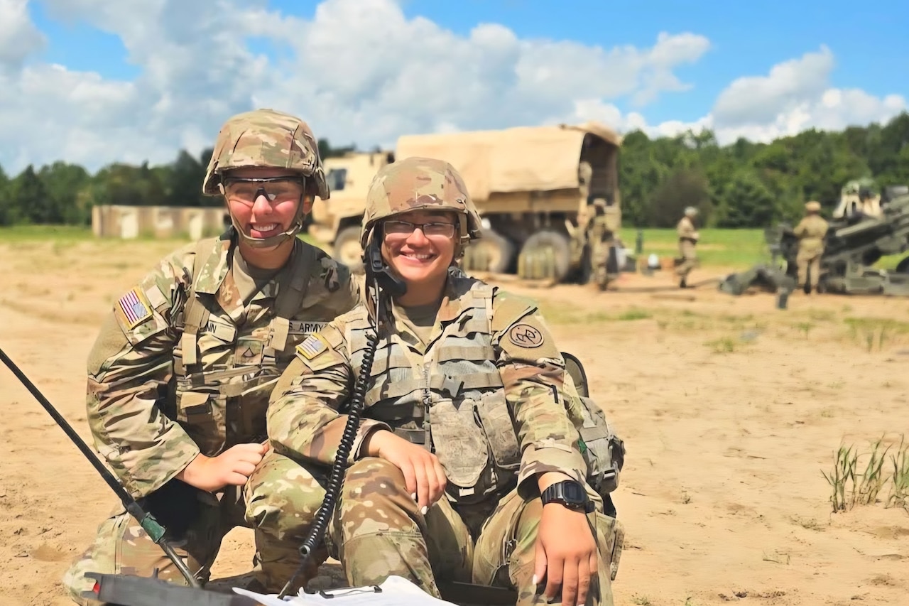 Two soldiers kneel and pose for a photo outside in front of a military vehicle and other soldiers during the day.