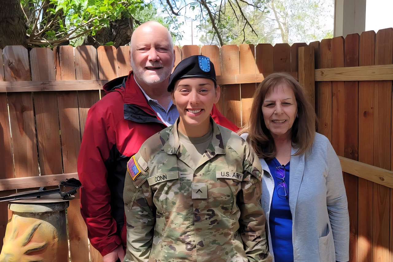 A service member poses for a photo with two people in civilian attire outside in front of a brown fence.
