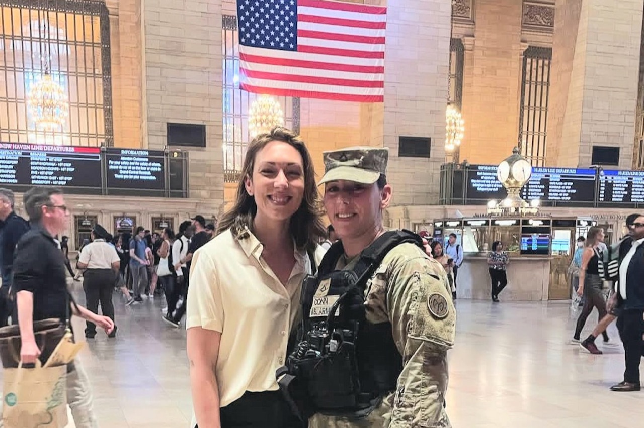 A woman in uniform poses next to a woman in civilian attire in the lobby of a train station.