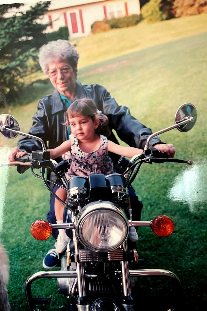 A toddler sits on a motorcycle with an elderly woman sitting behind holding the handlebars.