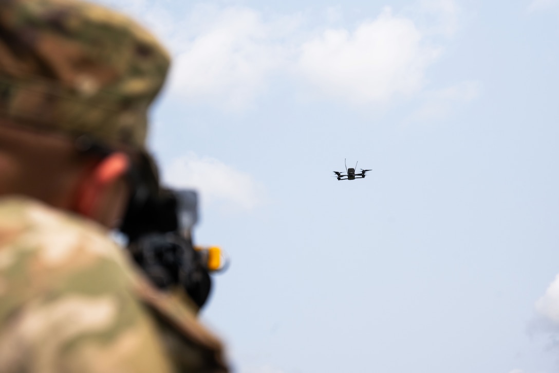 A soldier aims a rifle at an airborne drone with a blue sky and clouds in the background.