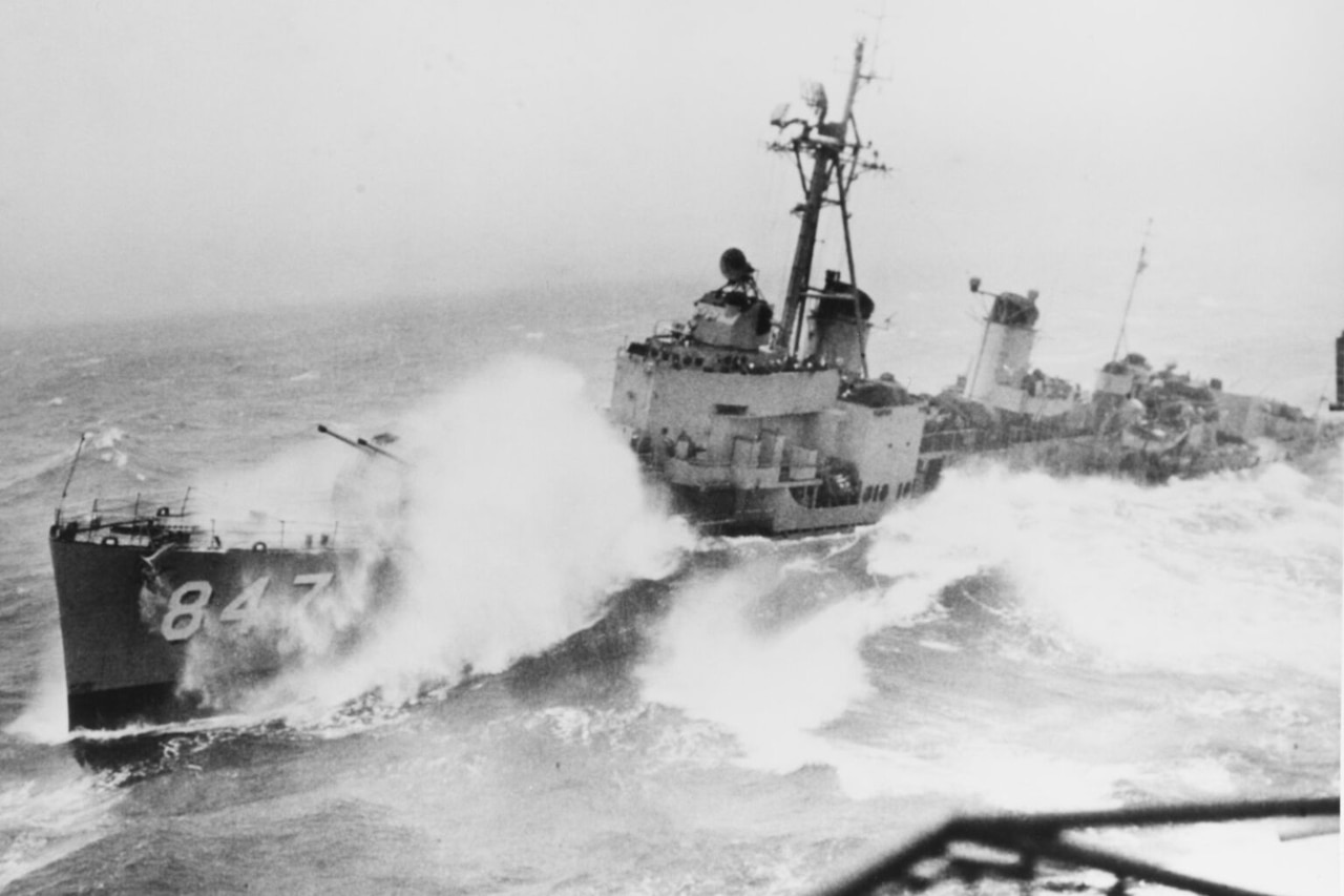 In a black and white photograph, a military vessel sails amid high, breaking waves.