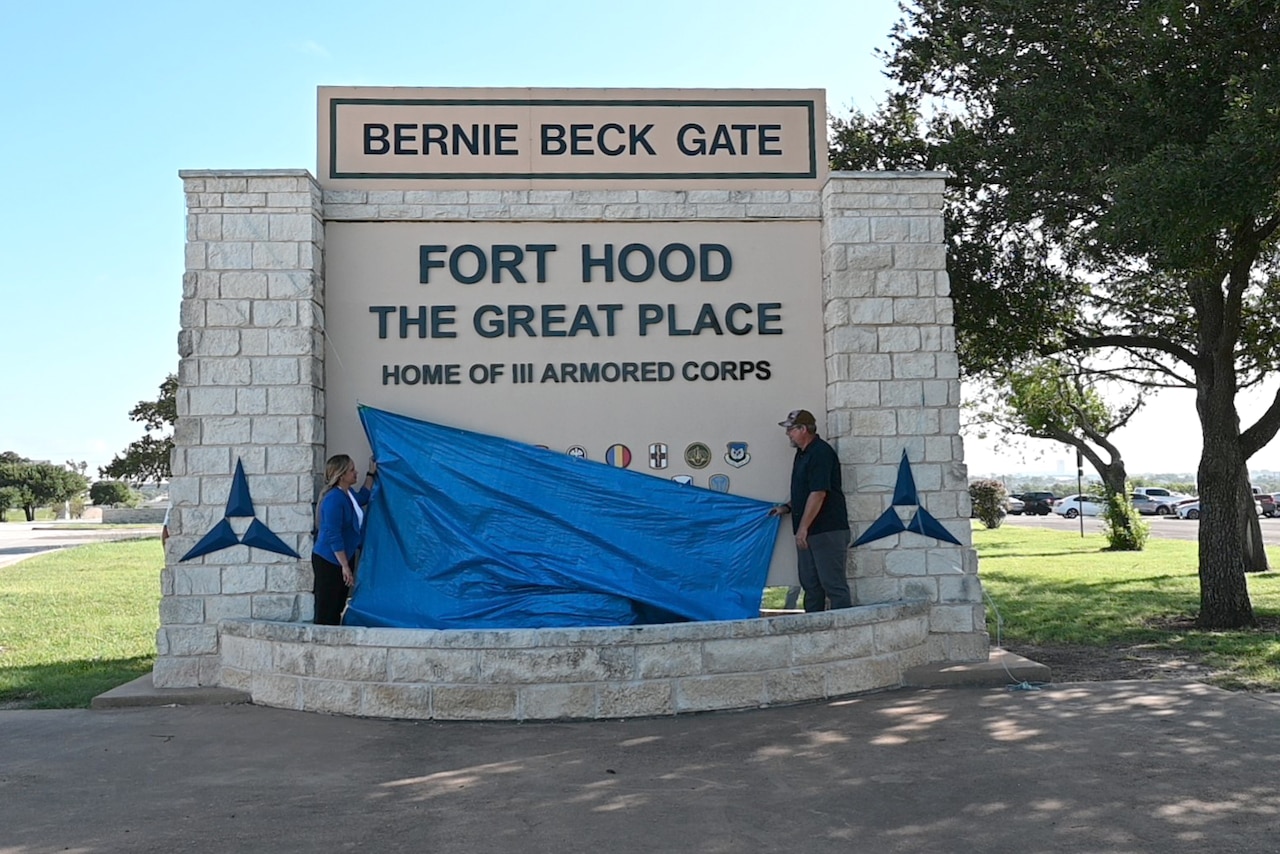 Workers remove a tarp from a sign, revealing the words "Fort Hood, the Great Place."