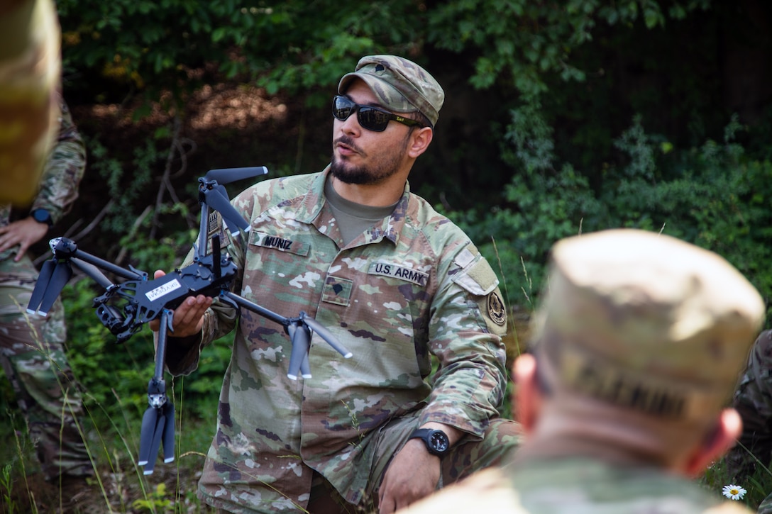 A soldier wearing sunglasses and holding a drone speaks to a group of other soldiers with green vegetation in the background.