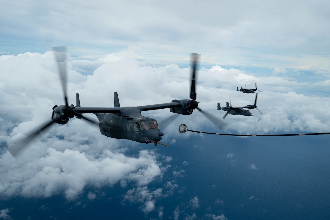 A tiltrotor aircraft prepares to connect with a fuel cable while flying over water; two other tiltrotor aircraft are nearby, with a cloudy sky in the background.