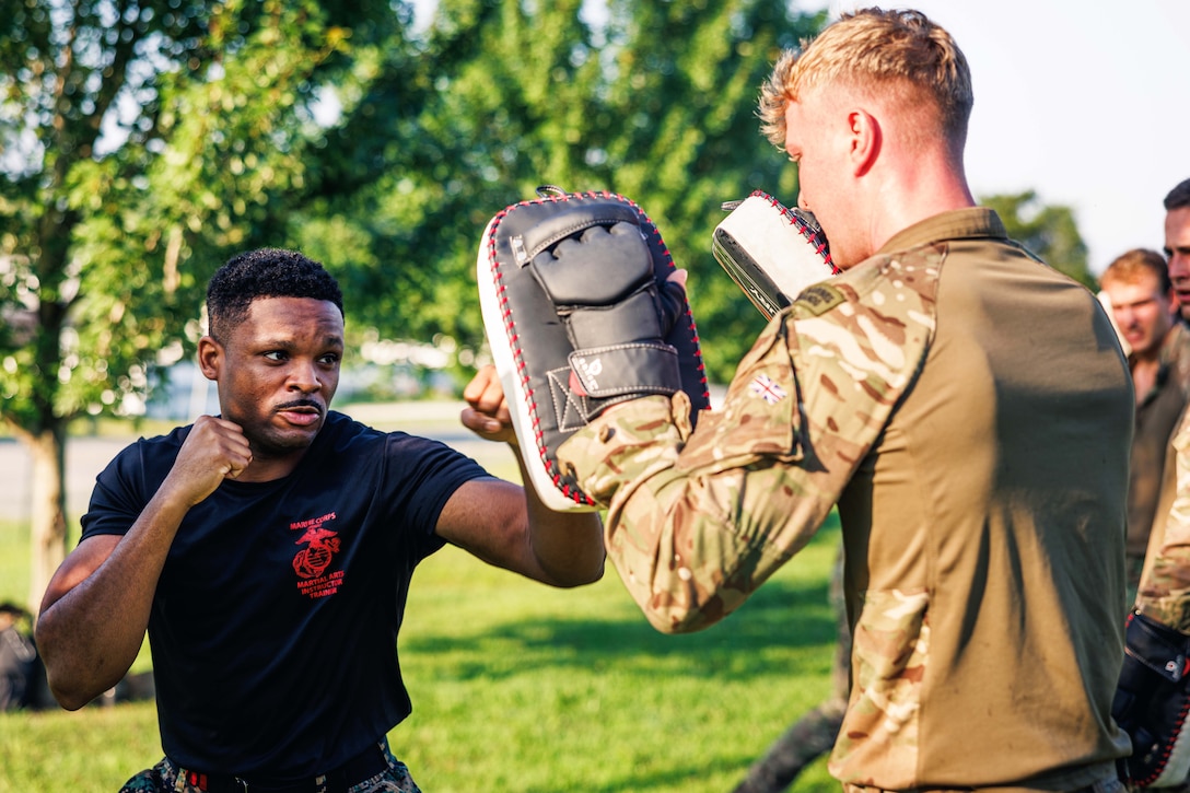 A U.S. Marine with fists raised prepares to punch the padded hands of a British marine in an outdoor setting as others look on, with trees and grass in the background.