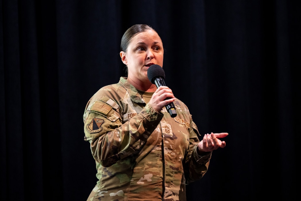 A woman, wearing military camouflage uniform, holds a microphone and gestures with her open hand