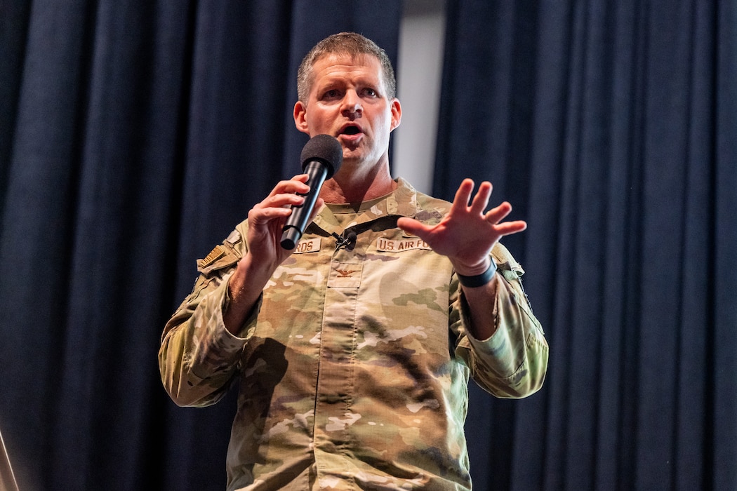 A man, wearing military camouflage uniform, holds a microphone and gestures with his open hand