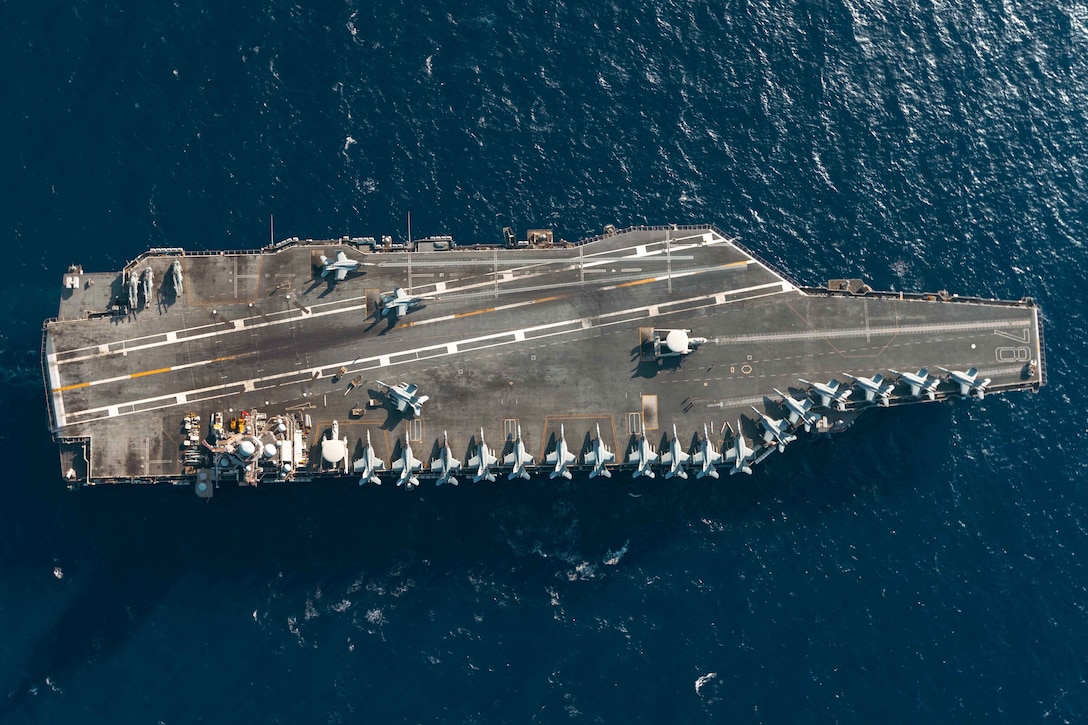 The deck of a ship at sea is shown from above, with fighter jets and other aircraft parked on it.