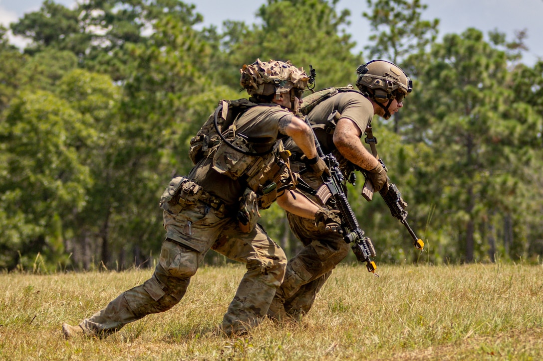 Two soldiers wearing helmets and carrying rifles move across a grassy field with trees in the background on a sunny day.