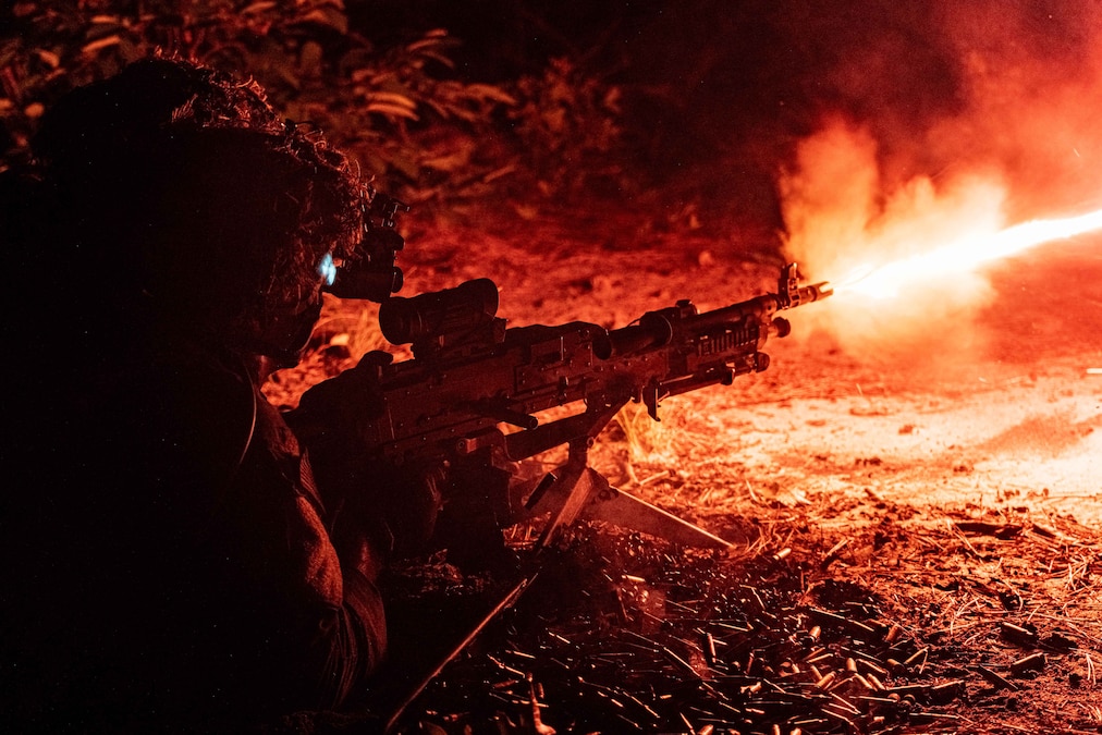 A soldier lying down fires a machine gun in dark conditions, leaving an orange trail from the barrel of the gun.