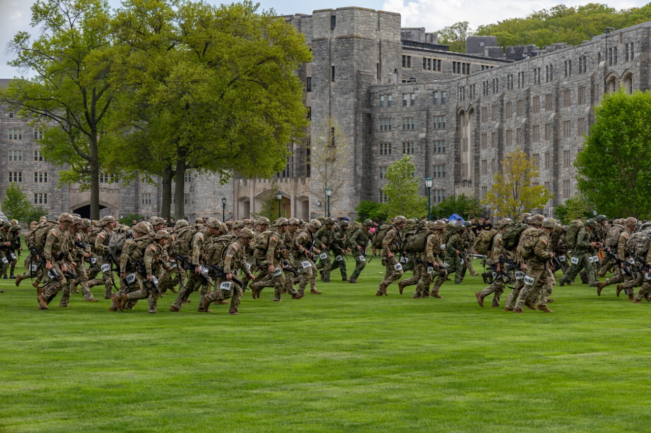 Dozens of uniformed personnel walk across a lawn in front of a stone building.