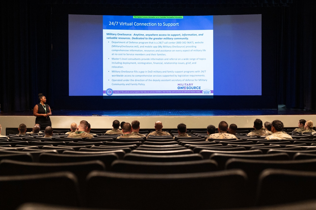 A group of people wearing camouflage military uniforms sits in an auditorium and listens as a woman in business attire stands at the front. A projection screen at the front of the room displays training slides.