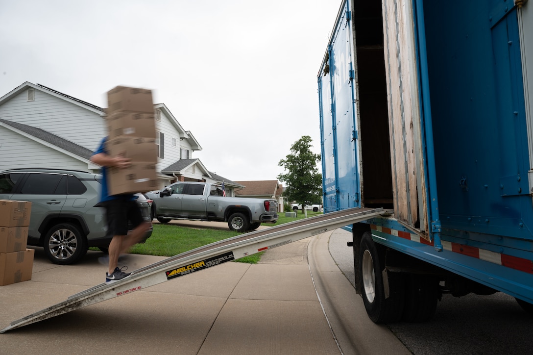 A person carries four cardboard boxes up a ramp to load onto a truck. A neighborhood is in the background.