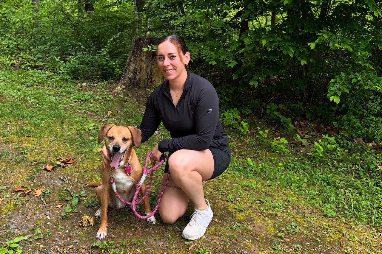 A woman sits with a dog on a walking trail in the woods.