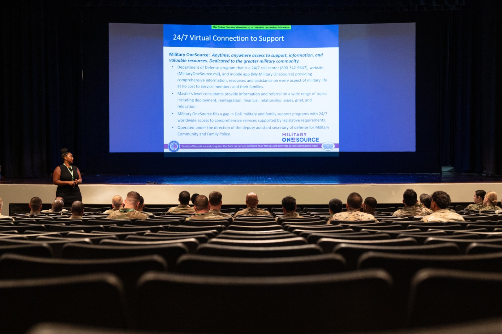A group of people wearing camouflage military uniforms sits in an auditorium and listens as a woman in business attire stands at the front. A projection screen at the front of the room displays training slides.