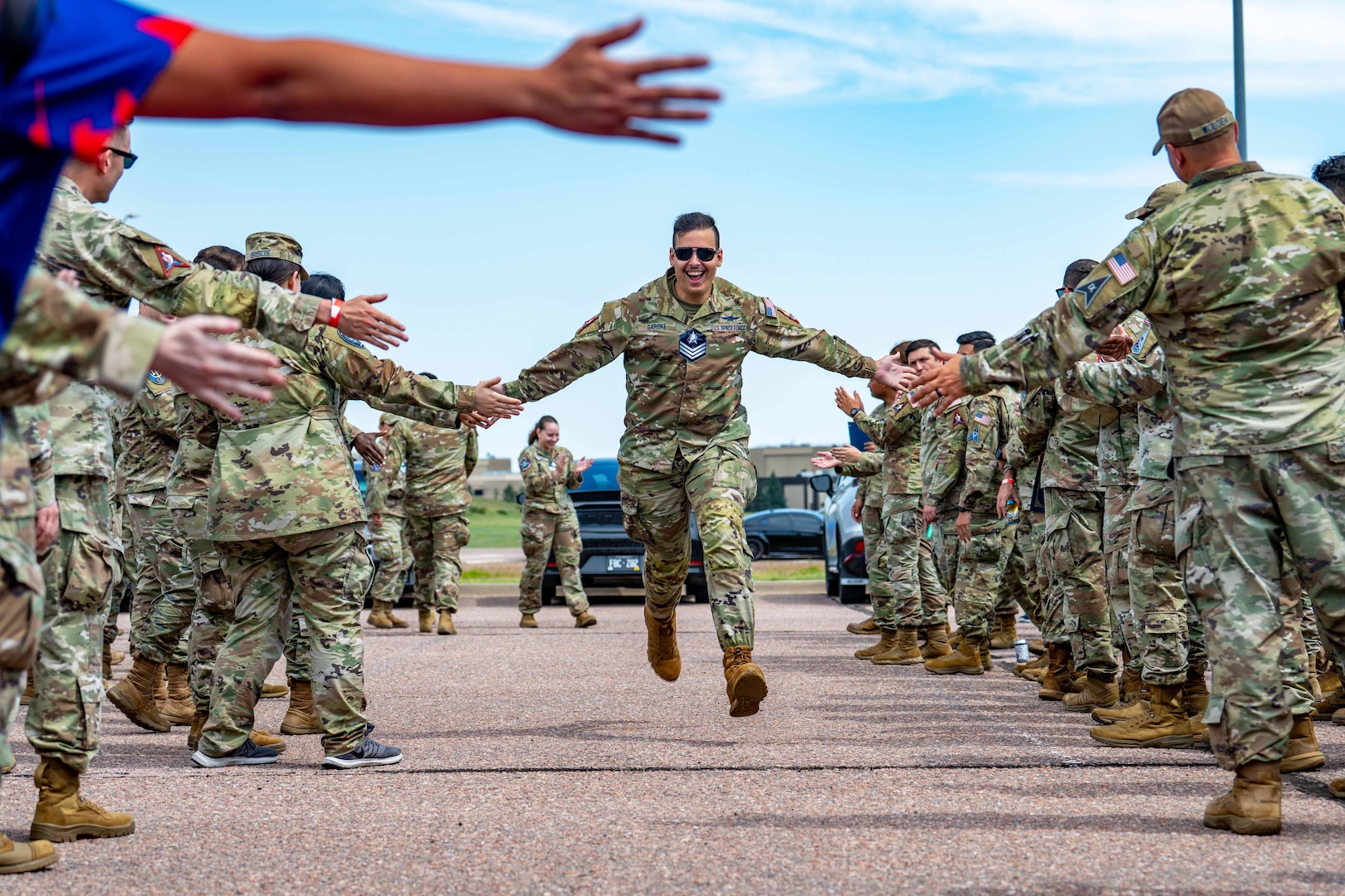 A guardian slaps hands with other guardians while running on a paved area with people on each side under a blue sky; vehicles are parked in the background.