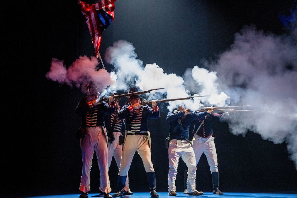 Five soldiers dressed in period uniforms fire weapons, creating clouds of smoke in a dark room.