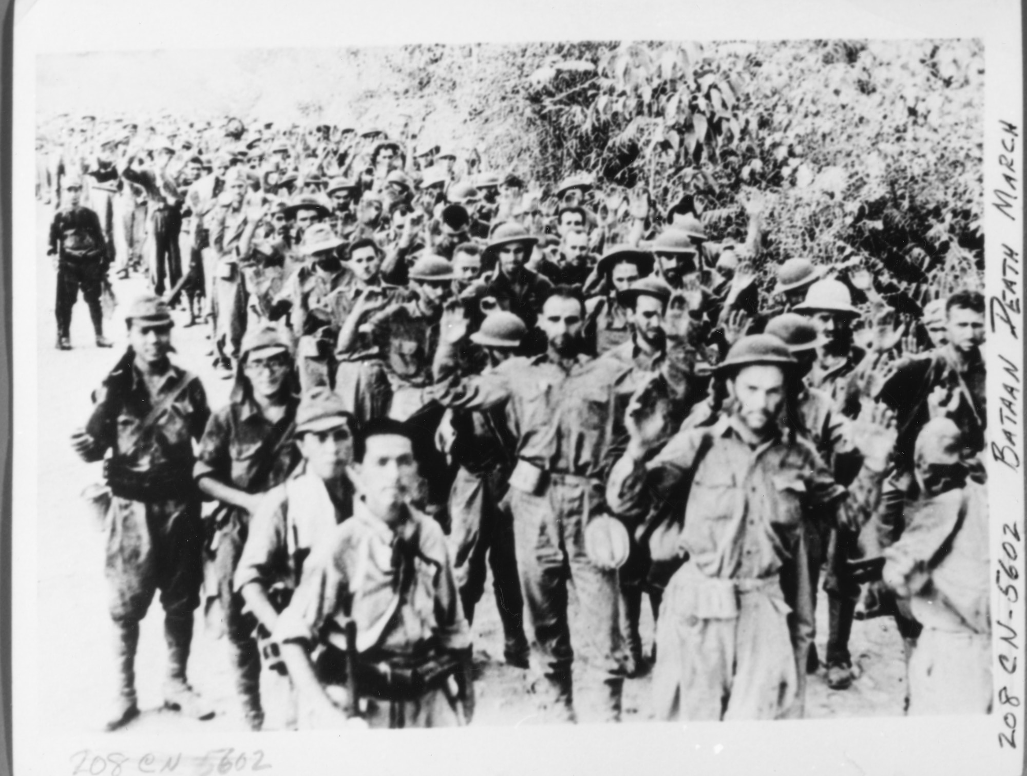 group of exhausted-looking men walking along a dirt road with guards
