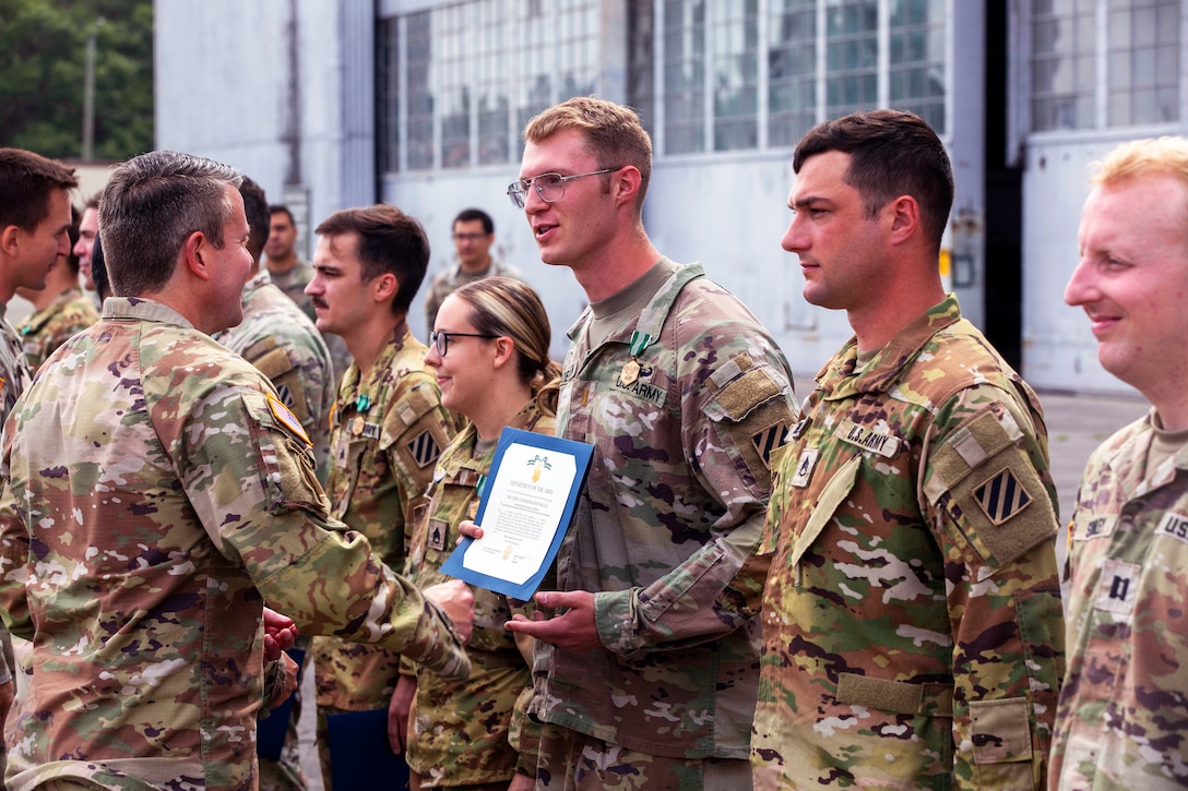 A soldier with a medal pinned to a collar receives a certificate from another soldier as others stand next to them in a row.